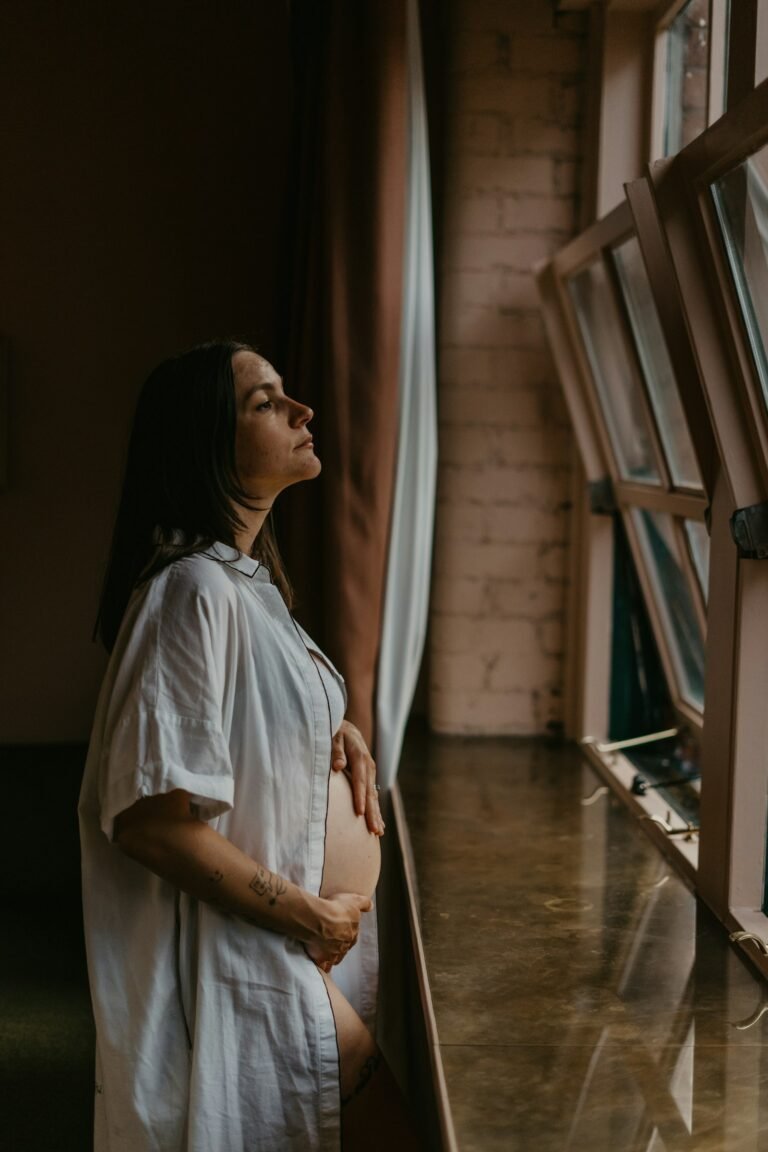 Gestante perto de uma janela, olhando para fora com a mão na barriga